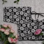 A black fabric garment with a repeating gray and white floral print, laid flat on a white textured surface with pink roses and green leaves around it.