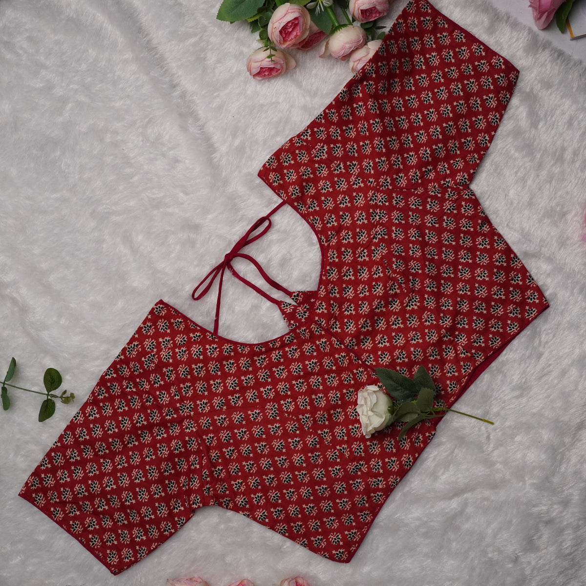 A red printed blouse top with a small repeating floral pattern, laid flat on a white textured fabric background with pink roses as decorative props.