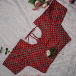 A red printed blouse top with a small repeating floral pattern, laid flat on a white textured fabric background with pink roses as decorative props.