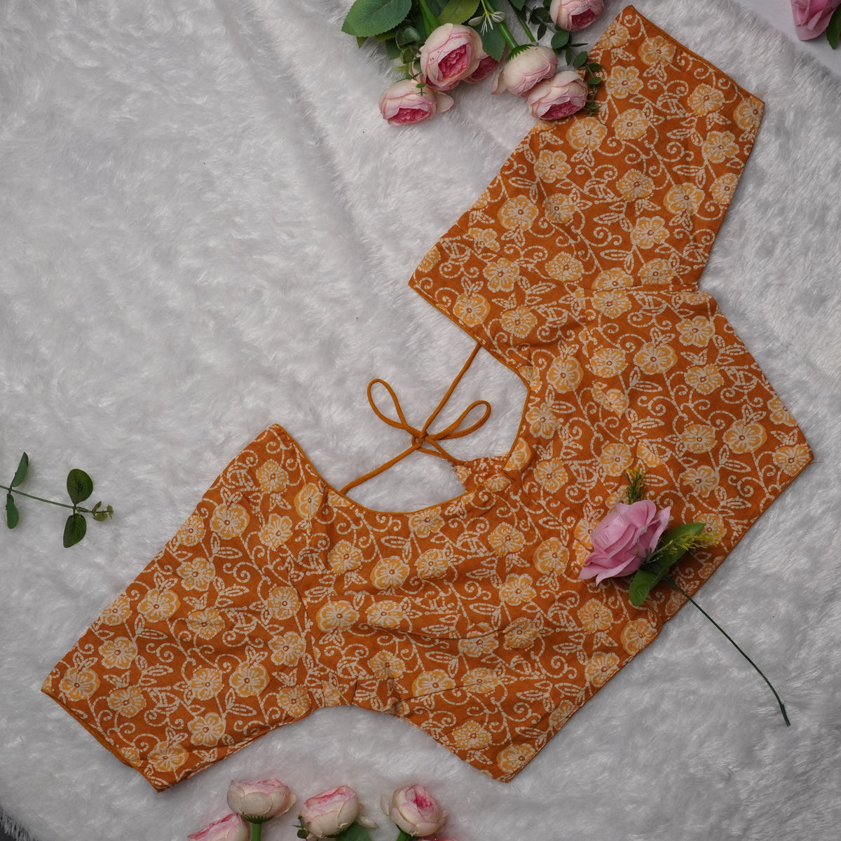 An orange and beige printed blouse piece with a tie neckline, laid flat on a white textured background with pink roses as decorative props.