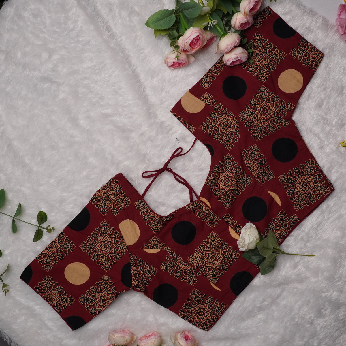 A flat-lay view of a traditional Indian blouse (sleeveless top) with a deep V-neckline, featuring a maroon base with black and gold geometric patterns and circular motifs, styled on a white textured fabric with pink roses.