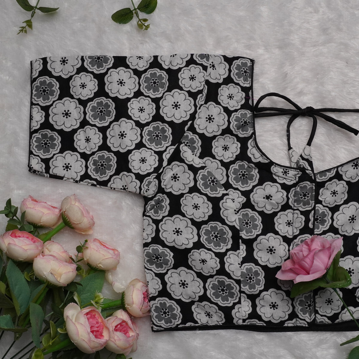 A black fabric garment with a repeating gray and white floral print, laid flat on a white textured surface with pink roses and green leaves around it.